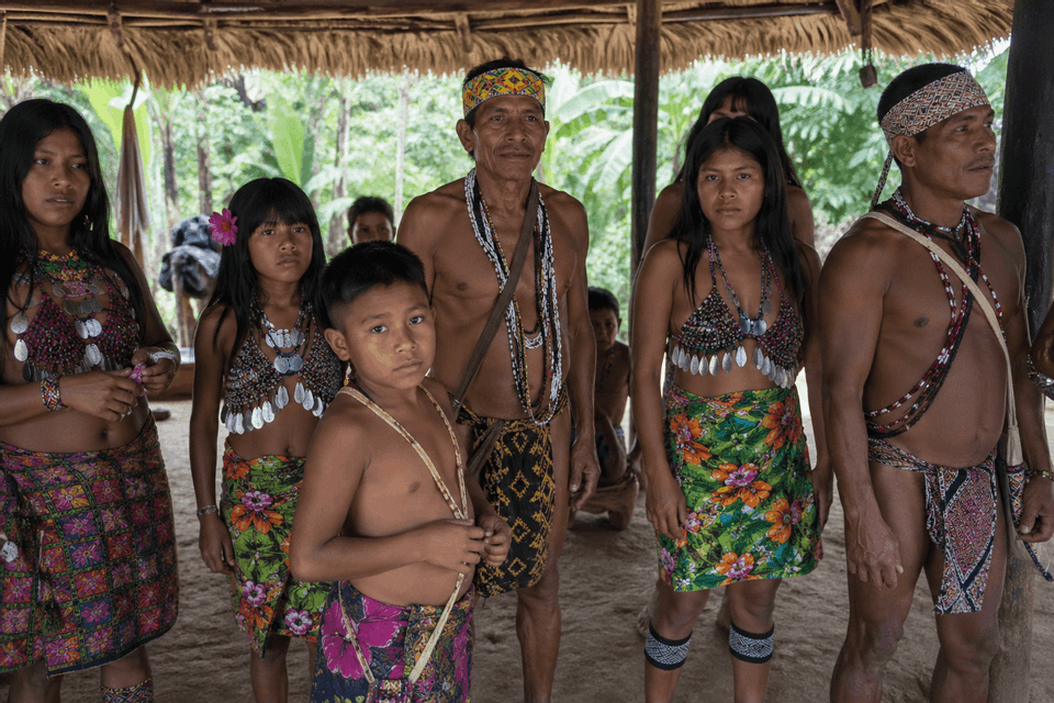 Un gruppo di adulti e bambini indigeni in abiti tradizionali si trova sotto un riparo di paglia in una giungla.