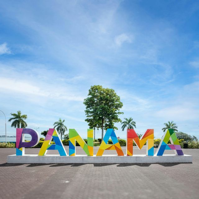 Un grande cartello colorato con la scritta 'PANAMA' si trova in una piazza con palme sotto un cielo blu parzialmente nuvoloso.