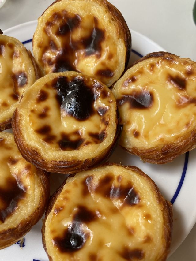 Un primo piano di alcuni pasteis de nata con croste friabili e la superficie caramellata su un piatto bianco.