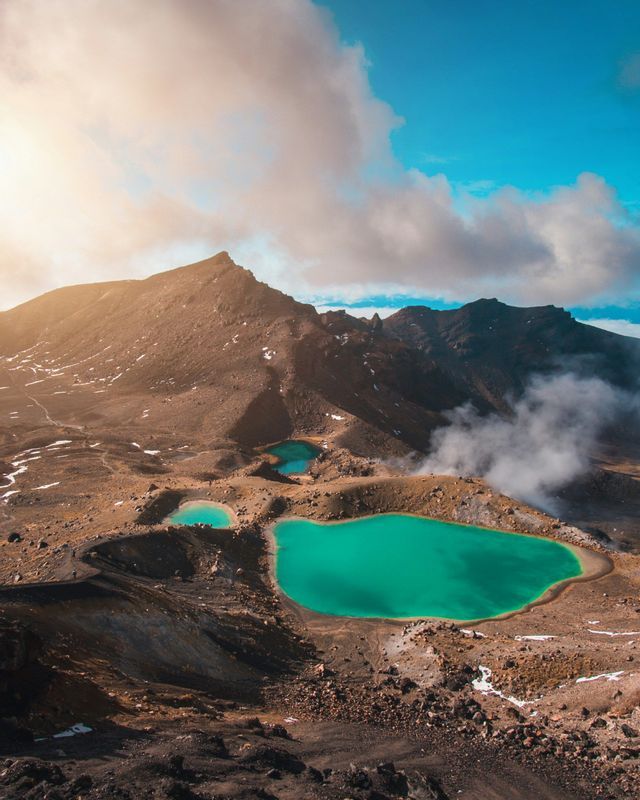 An aerial view of three turquoise crater lakes nestled in a rocky, volcanic landscape with steam rising from the ground.