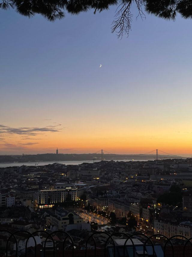 Un paesaggio urbano al crepuscolo visto dall'alto, con edifici illuminati, un ponte distante sull'acqua e una luna crescente nel cielo colorato.