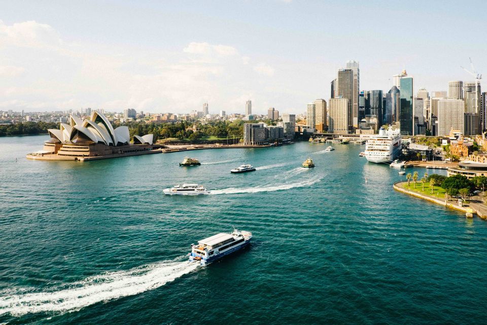 An aerial view of a busy harbor with ferries, a large opera house, and a modern city skyline under a clear sky.