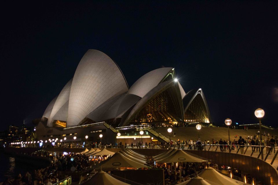 The Sydney Opera House illuminated at night, viewed from a crowded waterfront promenade with outdoor seating.