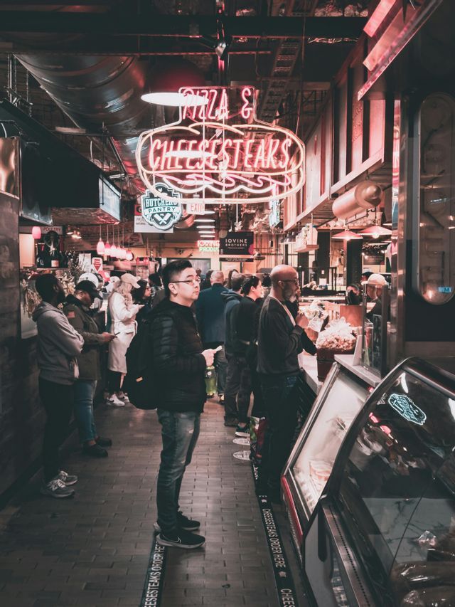 People waiting in line at an indoor food market under a large red 'Pizza & Cheesesteaks' neon sign.