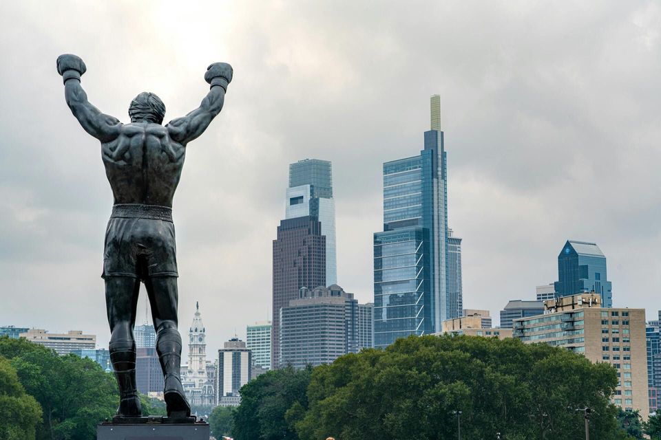 The back of a bronze statue of a boxer with raised arms in a victory pose, overlooking a modern city skyline under a cloudy sky.