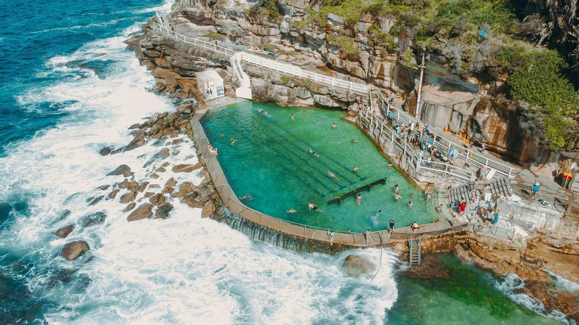 An aerial view of an ocean pool carved into a rocky cliff, with people swimming as waves crash against the barrier.