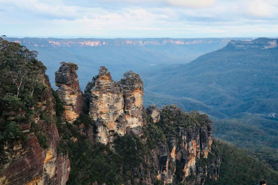 Three large rock pillars stand on a cliff edge overlooking a wide, forested mountain valley under a cloudy sky.