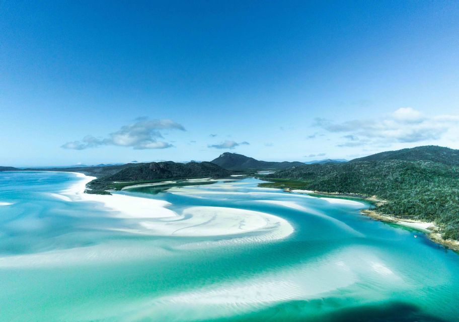 An aerial view of turquoise water swirling around white sandbars and green, hilly islands under a clear blue sky.