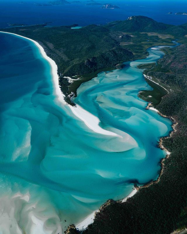 An aerial view of white sand swirling through vibrant turquoise water along a forested coastline, with distant islands on the horizon.