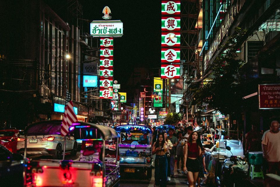 Una strada cittadina affollata di notte con persone che camminano e tuk-tuk nel traffico, illuminata da insegne al neon colorate con caratteri asiatici.