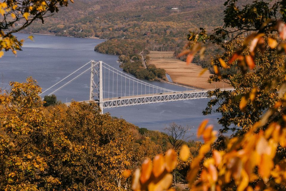 An aerial view of a white suspension bridge over a river, framed by trees with orange and yellow autumn leaves.