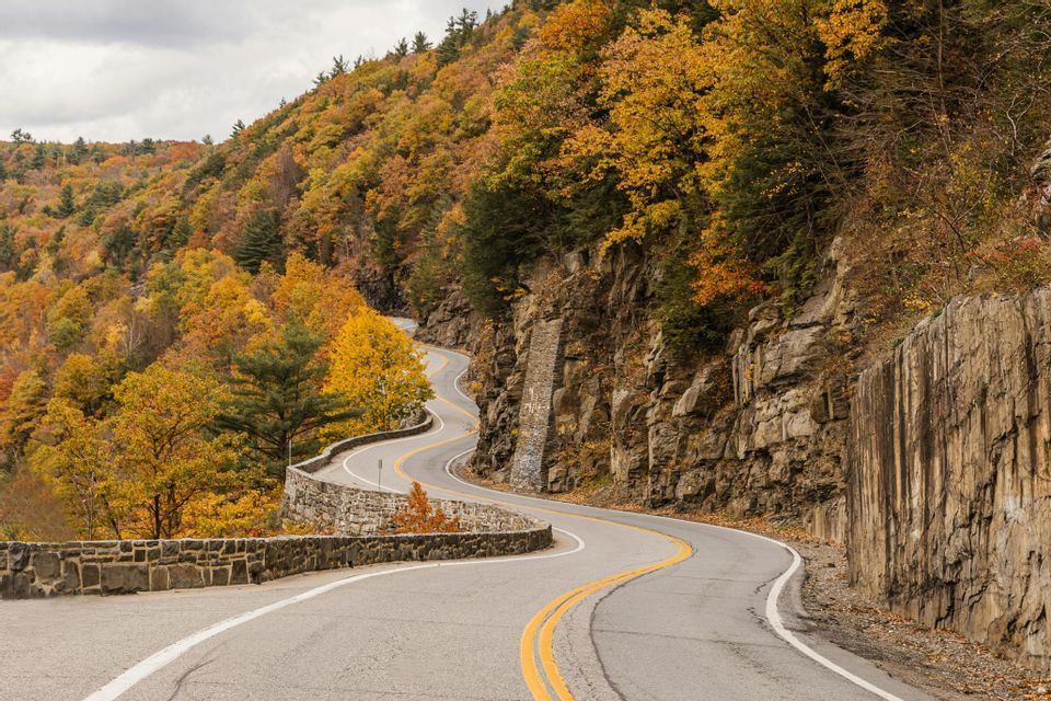A winding asphalt road curves through a rocky hillside covered in trees with yellow and orange autumn foliage.