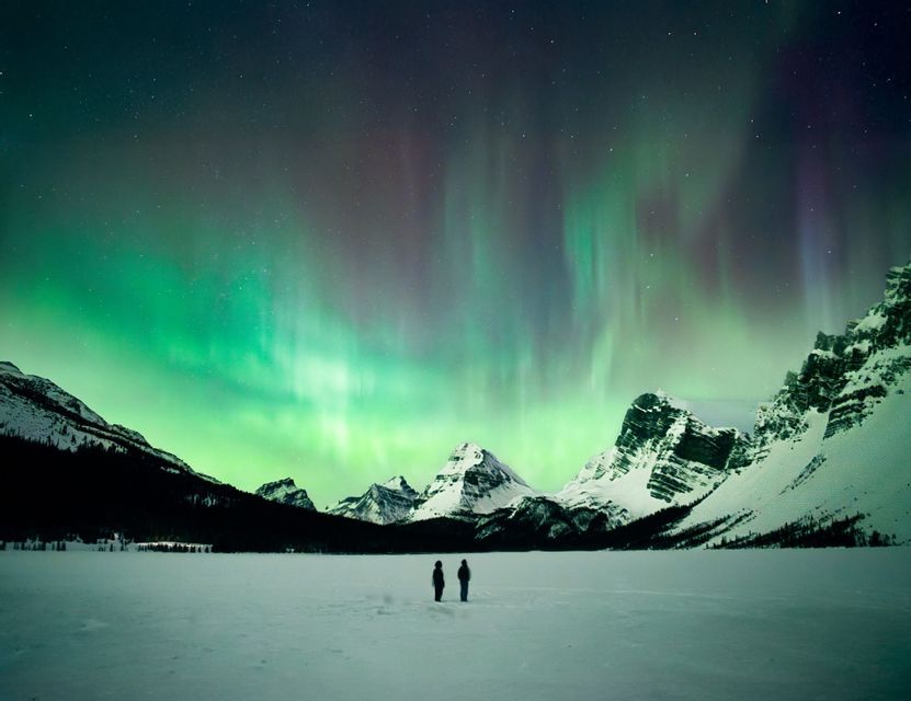 Two people stand on a vast snowy landscape watching the green and purple aurora borealis over snow-capped mountains.