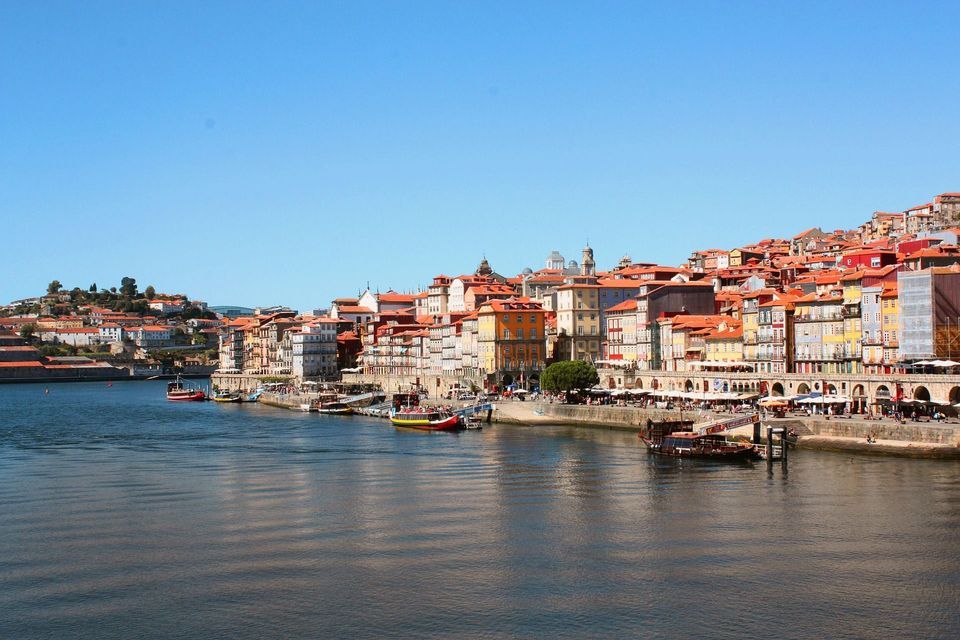 Edifici colorati su un pendio costeggiano un fiume, con barche ormeggiate lungo la passeggiata sul lungofiume sotto un cielo azzurro chiaro.
