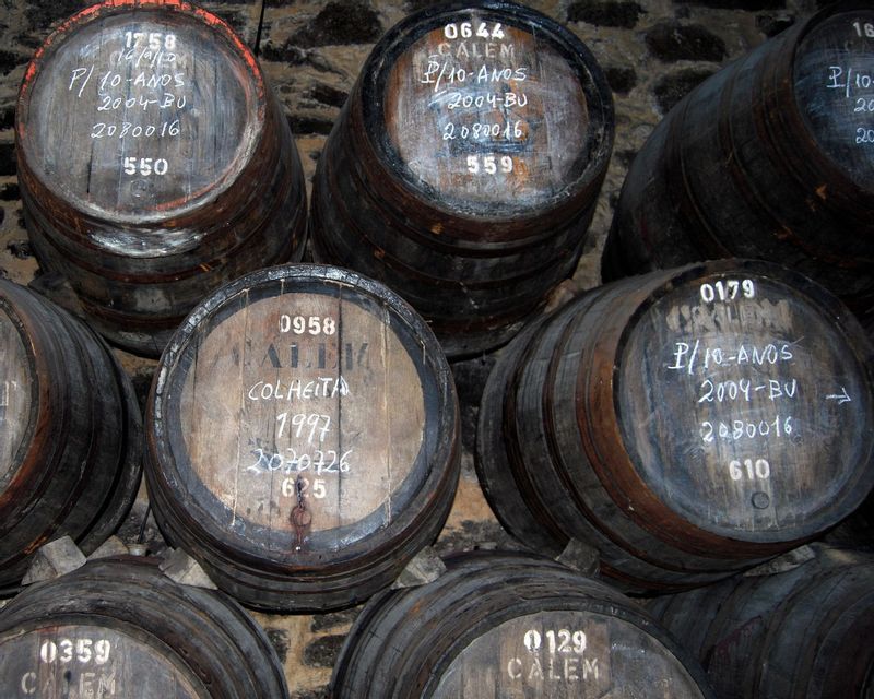 Botti di vino in legno impilate in una cantina di pietra, con scritte a gesso sulle estremità che indicano annata e contenuto.