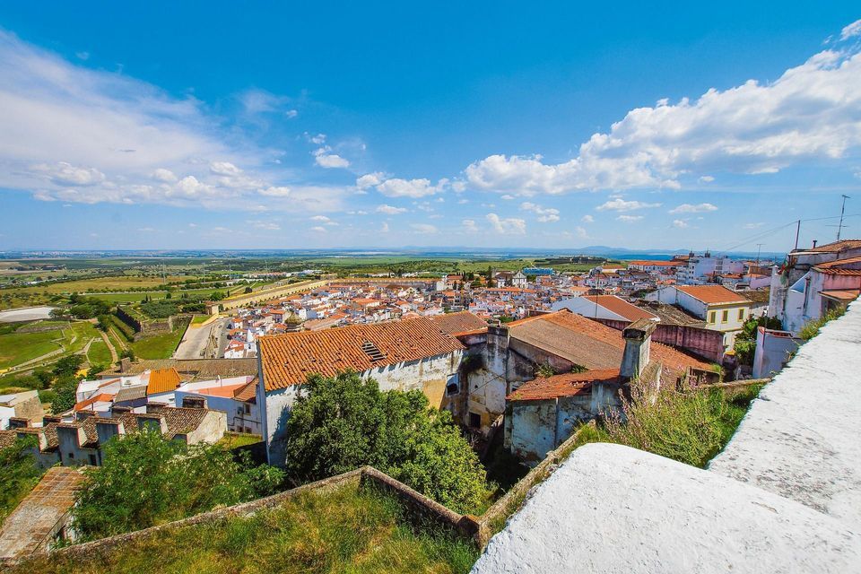 Veduta dall'alto di una cittadina con tetti di tegole rosse ed edifici bianchi, adagiata su un paesaggio verde sotto un cielo azzurro nuvoloso.