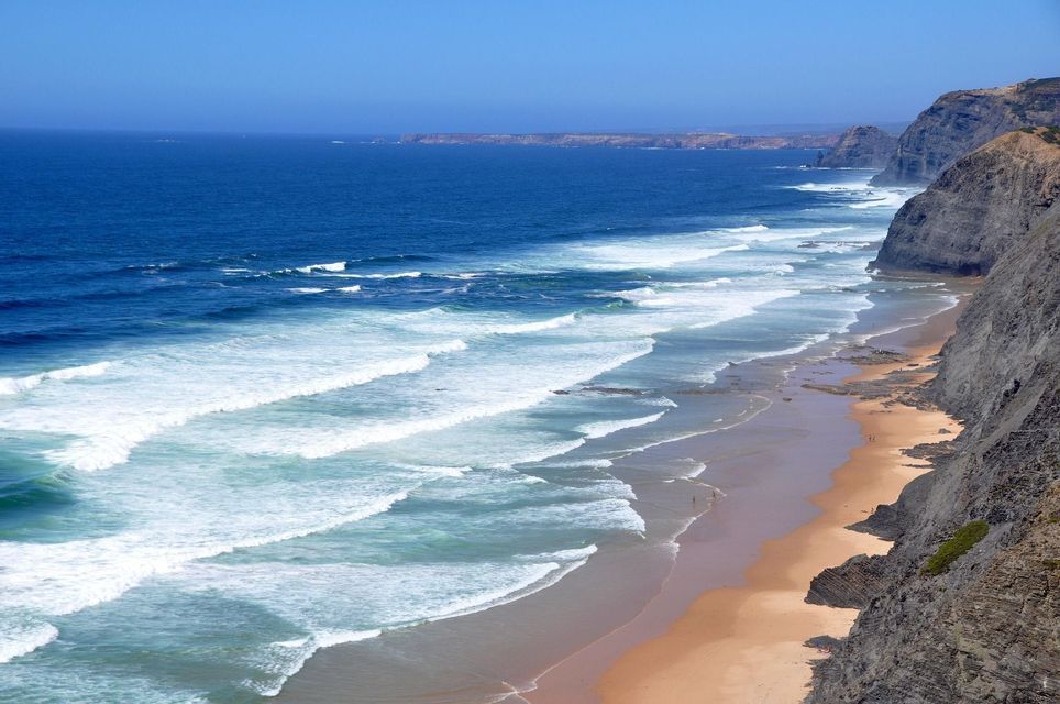 Onde bianche dell'oceano si infrangono su una spiaggia sabbiosa accanto a scogliere ripide e rocciose sotto un cielo azzurro e limpido.