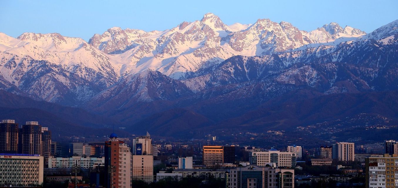 Eine Stadtlandschaft mit Gebäuden liegt am Fuße eines weitläufigen, schneebedeckten Gebirges, das von Sonnenlicht unter klarem Himmel beleuchtet wird.