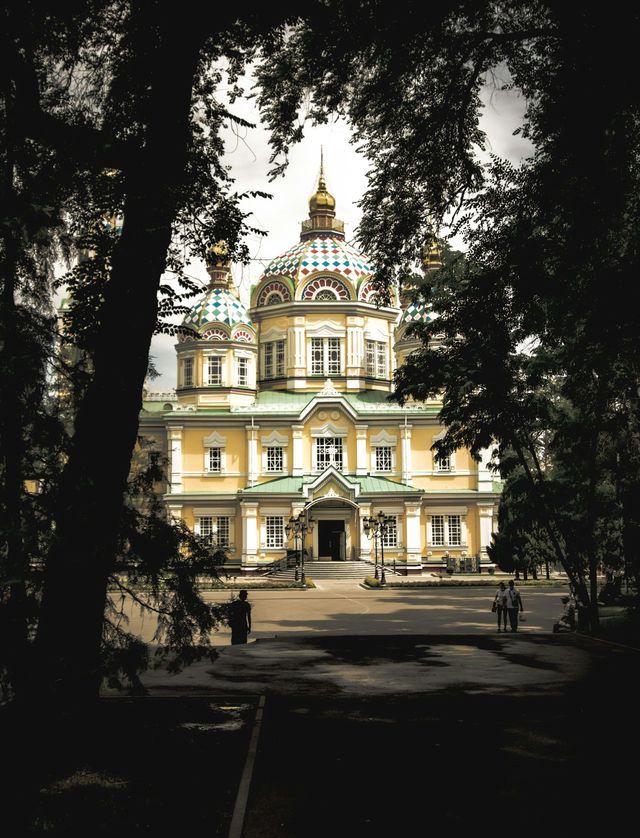 Eine gelbe orthodoxe Kathedrale mit farbenfrohen Kuppeln und goldenen Spitzen, aus der Ferne durch einen Rahmen dunkler Bäume betrachtet.