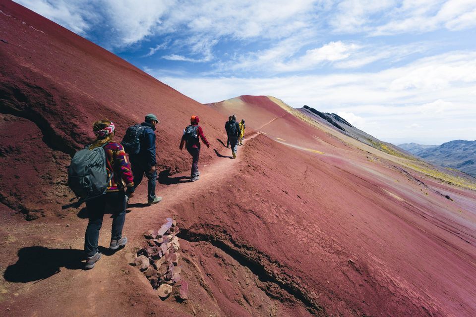 Un viaggio di gruppo WeRoad che fa trekking in fila indiana lungo uno stretto sentiero sul fianco di una vasta montagna rossa sotto un cielo parzialmente nuvoloso.