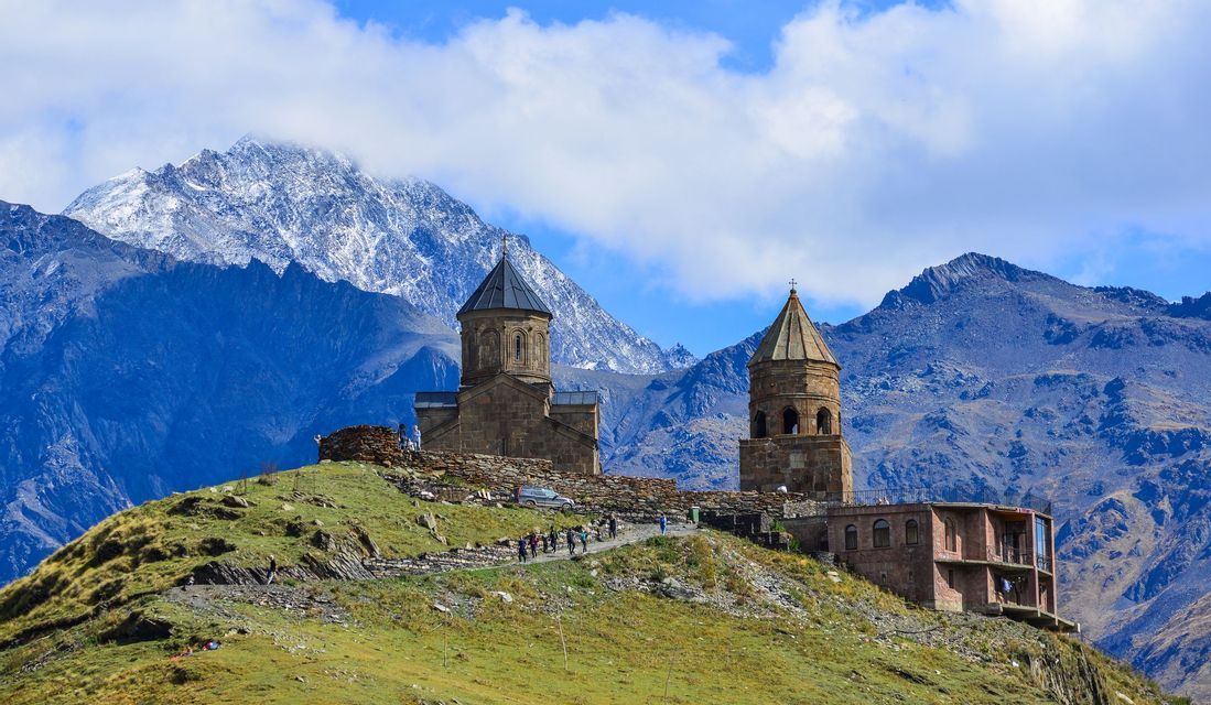 Un voyage en groupe WeRoad sur un chemin menant à un monastère de pierre, sur une colline verdoyante, avec de grandes montagnes enneigées en arrière-plan.
