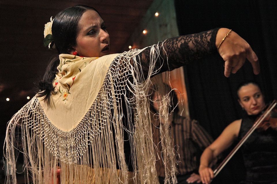 Une danseuse de flamenco portant un châle à franges et des manches en dentelle se produit sur scène avec un violoniste en arrière-plan.