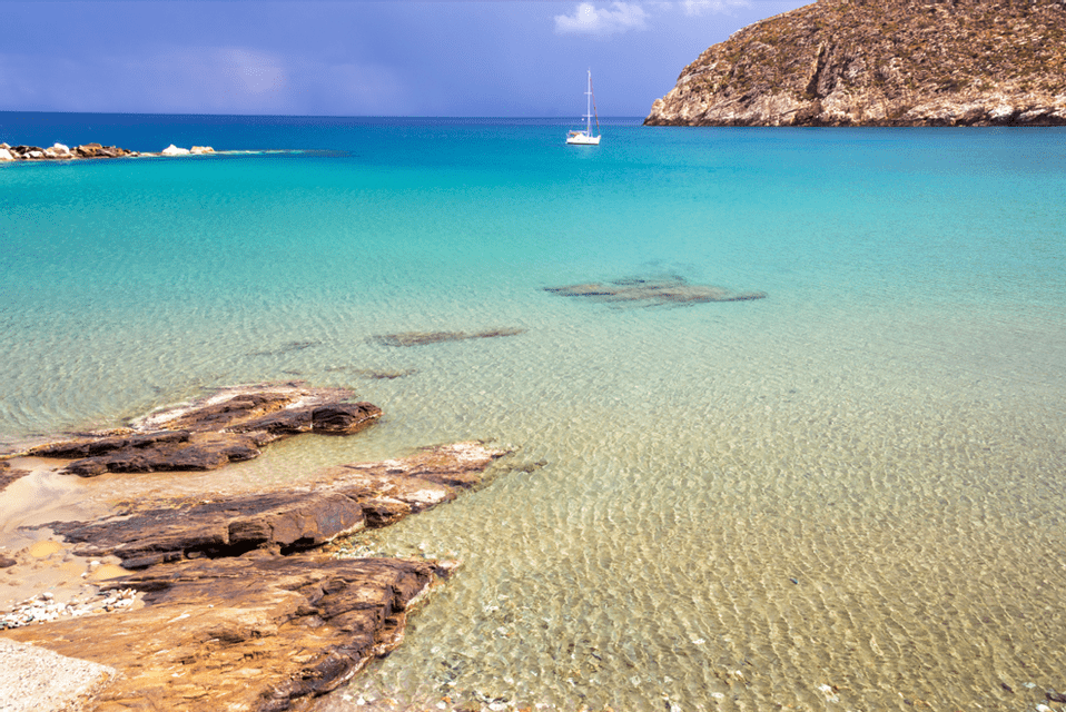 Una barca a vela bianca galleggia in una baia con acqua limpida e turchese, una costa rocciosa in primo piano e una costa collinare in lontananza.