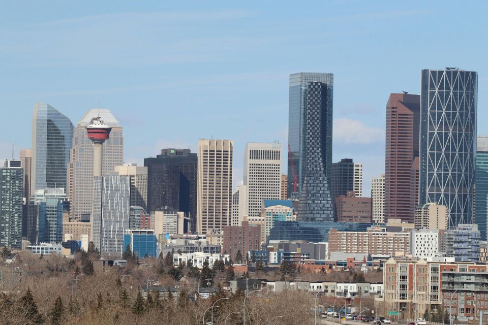 A dense city skyline featuring numerous skyscrapers and a tall observation tower under a pale blue sky, with trees in the foreground.