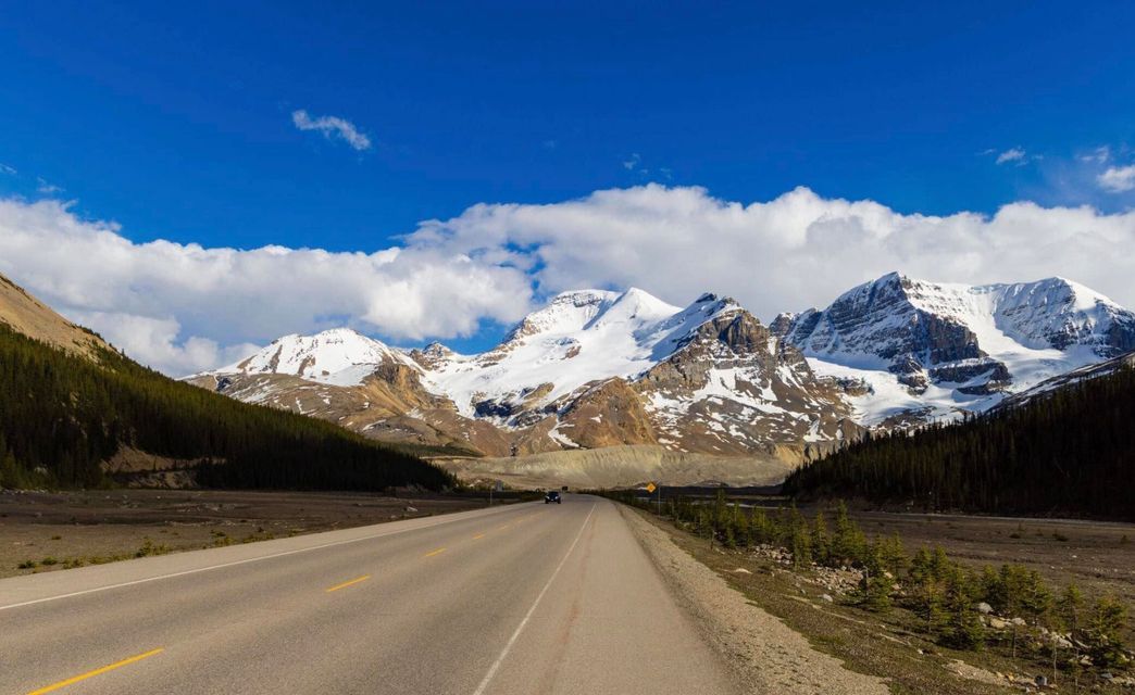 A paved road runs through a valley towards a range of towering, snow-capped mountains under a bright blue sky.