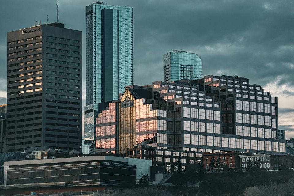 A cityscape of modern skyscrapers with glass facades reflecting the sunset under a dark, cloudy sky.