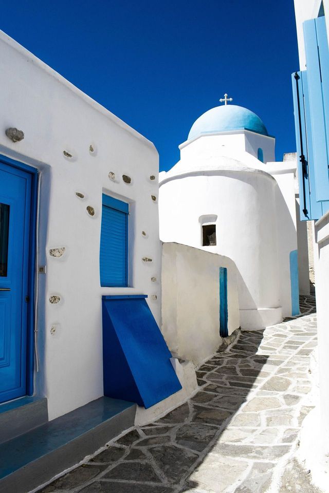 Un angusto vicolo in pietra tra edifici imbiancati a calce con porte blu, che conduce a una chiesa dalla cupola azzurra sotto un cielo limpido.