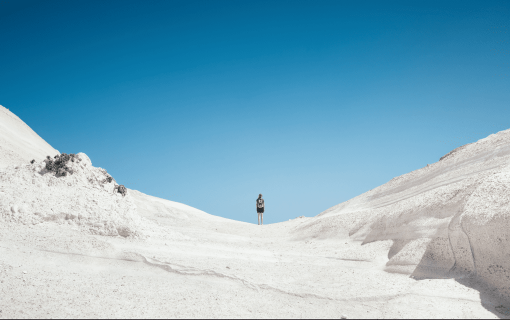 Un viandante solitario con uno zaino si staglia su una duna bianca contro un cielo blu intenso.