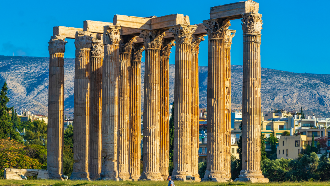 Alte colonne corinzie baciate dal sole di un'antica rovina, con una città e montagne visibili sullo sfondo sotto un cielo blu.