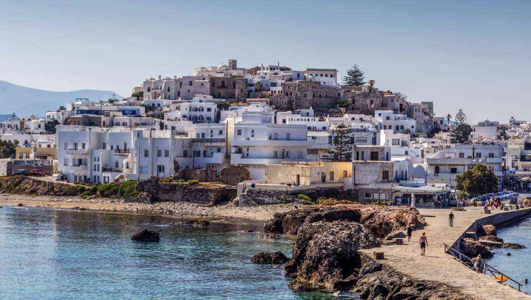 Un borgo collinare di case bianche si affaccia su un mare calmo, con persone che passeggiano lungo un molo di pietra.