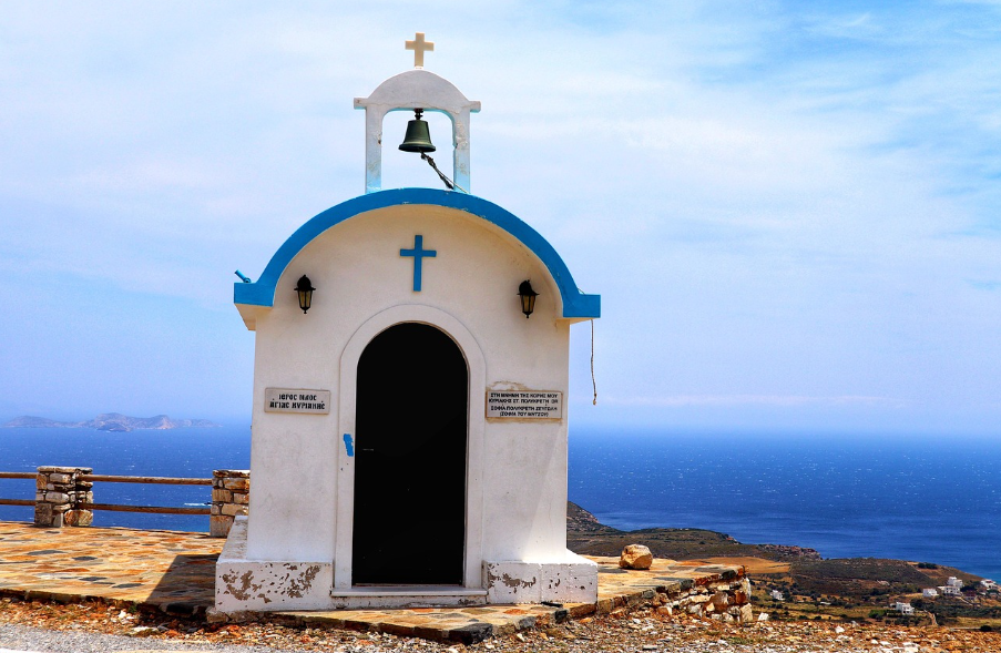 Una piccola cappella imbiancata, con dettagli blu e un campanile, si erge su una scogliera a picco sul mare azzurro.