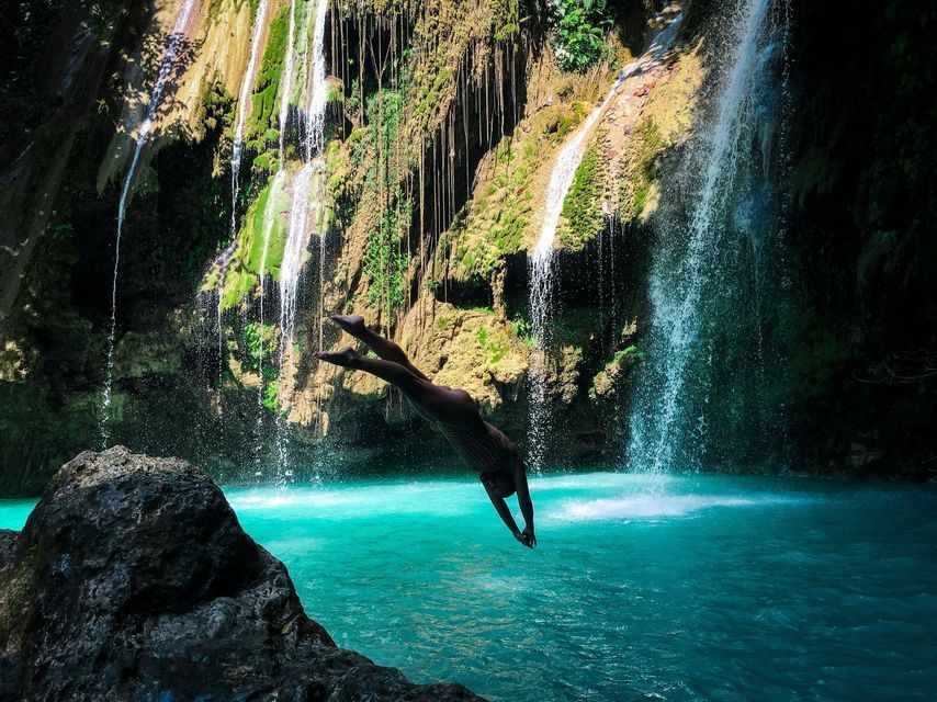A person dives headfirst from a rock into a turquoise pool of water at the base of lush, cascading waterfalls.