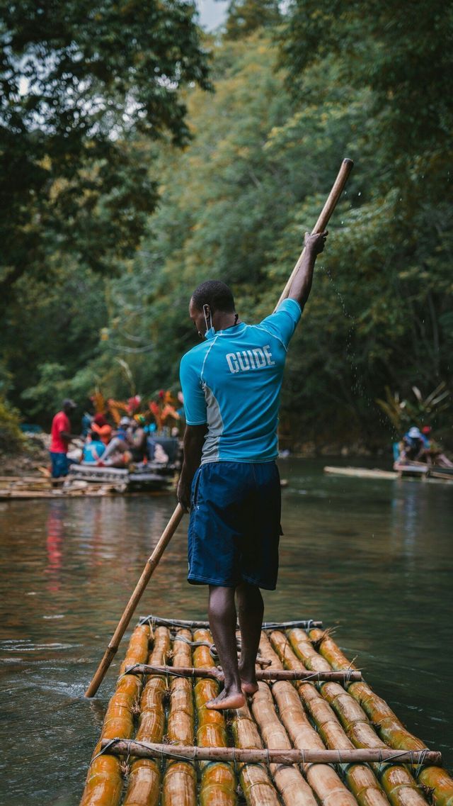 A guide seen from behind stands on a bamboo raft, using a long pole to navigate a river with a WeRoad group trip in the background.