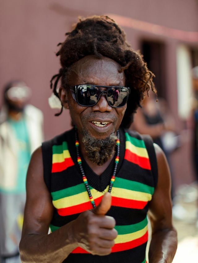 A man with dreadlocks and sunglasses smiles and gives a thumbs-up while wearing a striped vest.