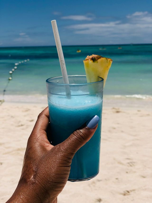 A hand with blue nail polish holds a frosty blue cocktail with a pineapple slice on a white sand beach.