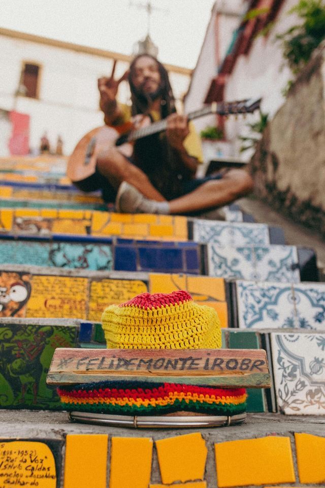 A man with dreadlocks plays guitar and makes a peace sign while sitting on colorful, tiled mosaic stairs, with a hat in the foreground.