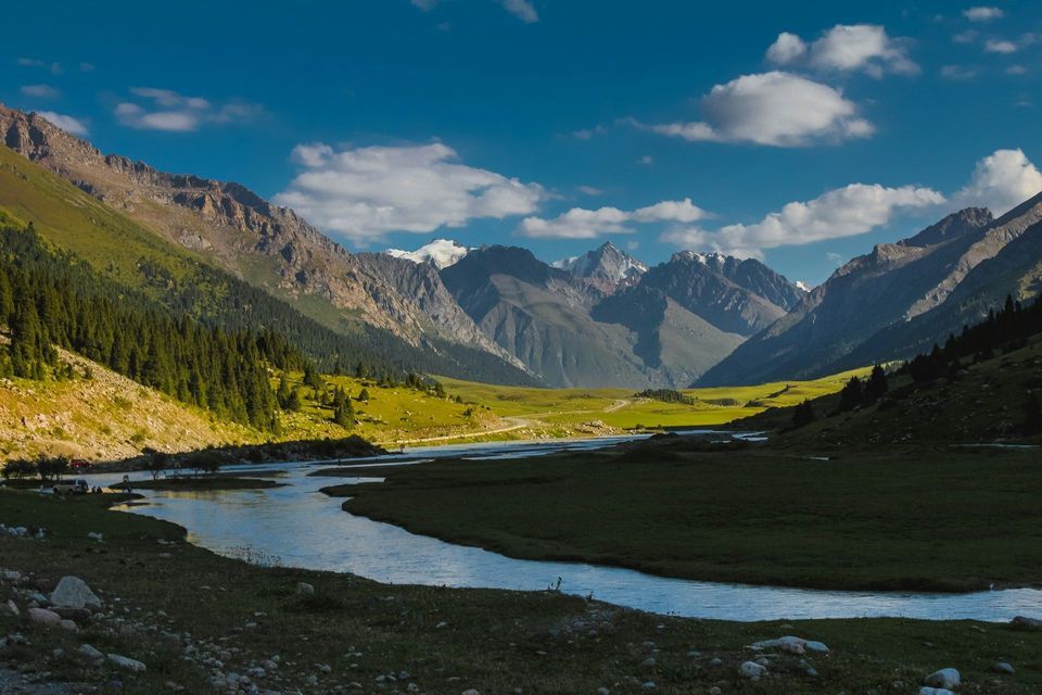 Ein Fluss schlängelt sich durch ein üppig grünes Tal auf eine entfernte Kette schneebedeckter Berge zu, wo eine WeRoad-Gruppe am Ufer rastet.