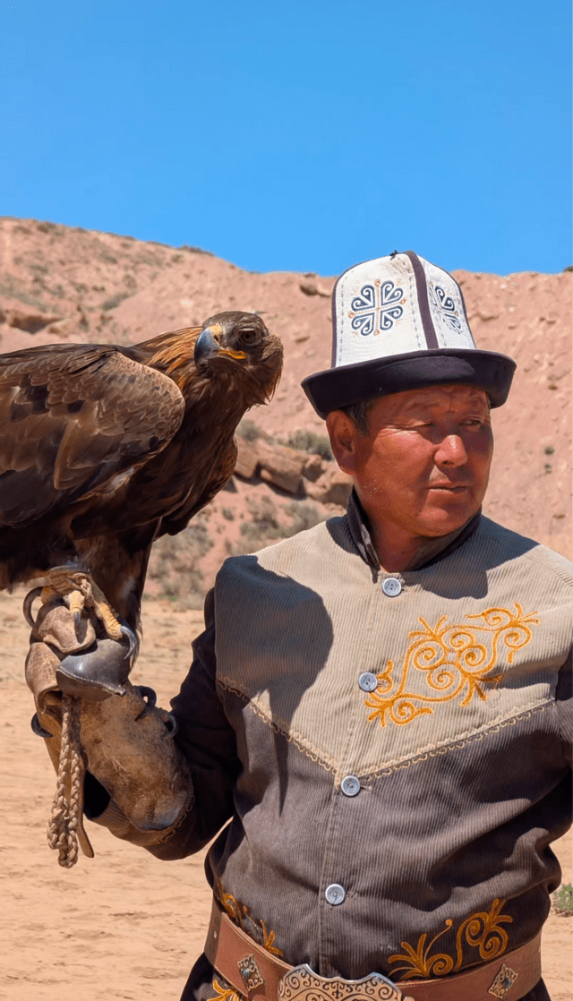 Ein Mann in traditioneller Kleidung hält einen Steinadler auf seiner behandschuhten Hand in einer hügeligen, trockenen Landschaft unter einem klaren blauen Himmel.