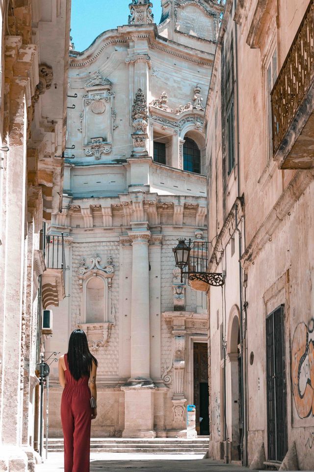 Una donna in tuta rossa vista di spalle, in piedi in uno stretto vicolo di pietra che guarda un ornato edificio barocco.