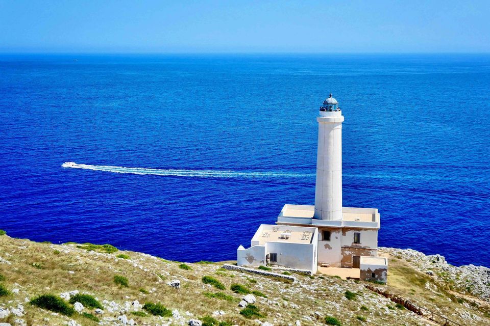 Un faro bianco si erge su una scogliera rocciosa a picco su un mare blu profondo, mentre un motoscafo sfreccia.