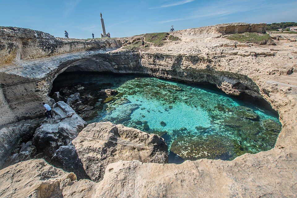 Una piscina naturale di acqua cristallina turchese all'interno di una caletta rocciosa, con persone che camminano sulle scogliere soprastanti sotto un cielo azzurro.