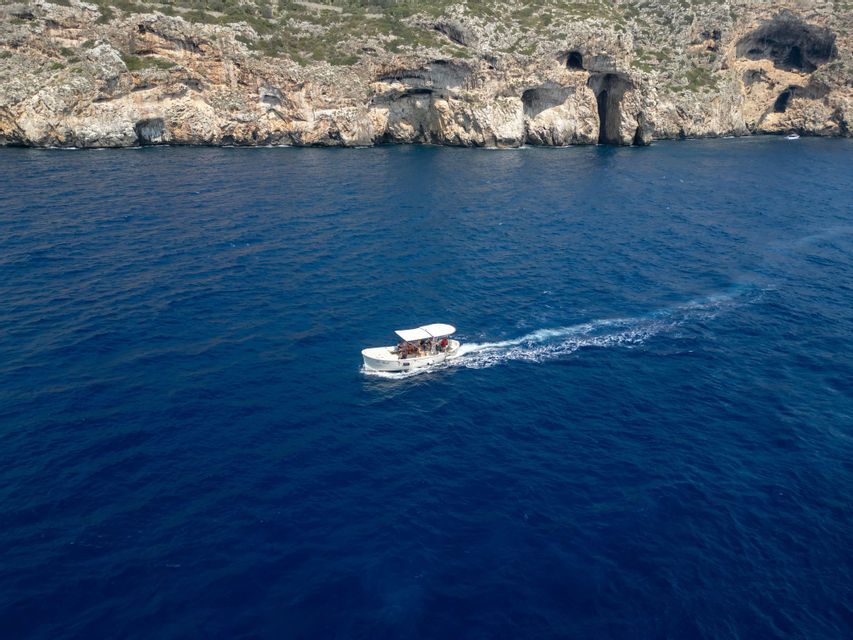 Una veduta aerea di un viaggio di gruppo WeRoad su una barca bianca che naviga in acque blu profonde verso una scogliera rocciosa con grotte marine.