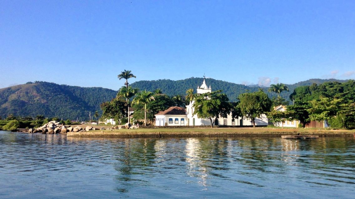 Eine weiße Kolonialkirche an einer üppigen Küste spiegelt sich im Wasser, mit grünen Hügeln im Hintergrund unter klarem Himmel.