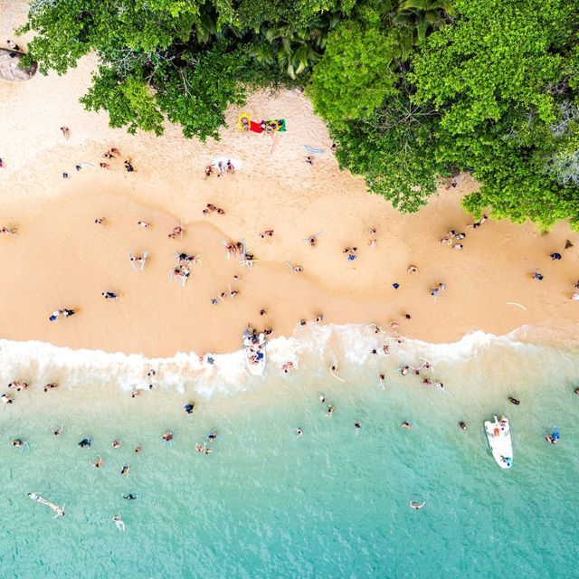 Luftaufnahme eines belebten Strandes mit Menschen, die im türkisfarbenen Wasser schwimmen und am goldenen Sandstrand entspannen.