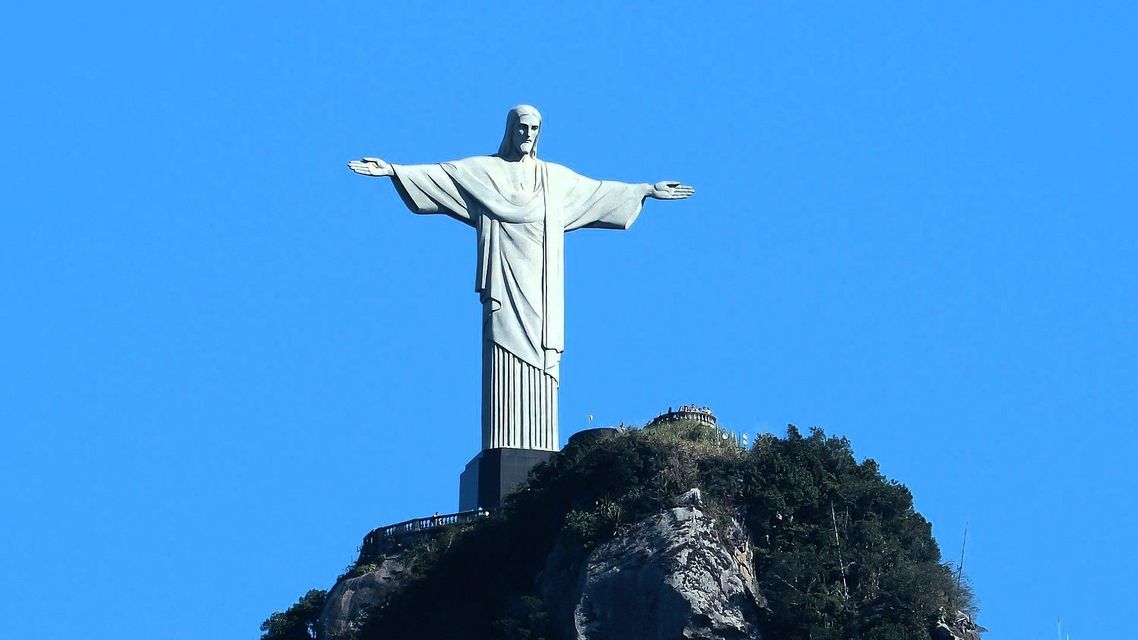 Eine imposante Männerstatue ragt mit ausgebreiteten Armen auf einem felsigen Berggipfel unter klarem blauem Himmel empor.