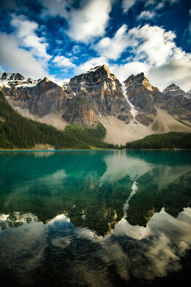 A rocky, snow-capped mountain range reflects onto the calm, turquoise water of a lake, with a pine forest on the shore.
